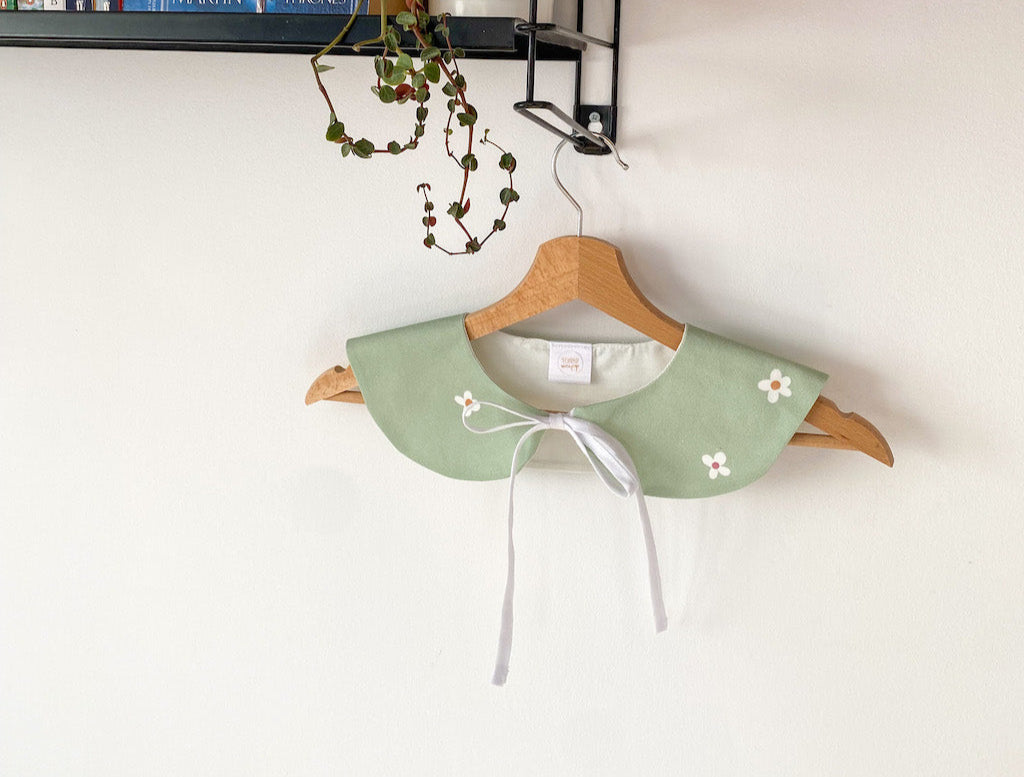 Green collar with floral details on a wooden hanger against a white wall with books and decorative items.