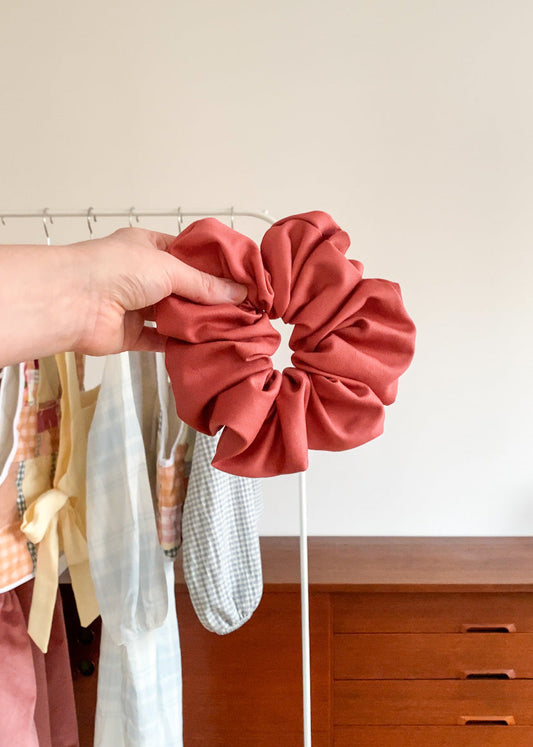 An oversized pink cotton twill scrunchie held in a person's hand, with a blurred background featuring various clothing items and a wooden hanger.