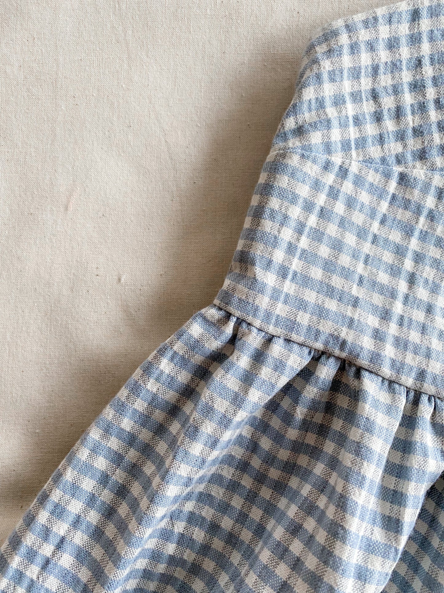 Close-up picture of the Alice dress, a poofy midi length dress made from lightweight deadstock cotton in a blue and white gingham print. The dress is sleeveless and ties into bows at the shoulders.