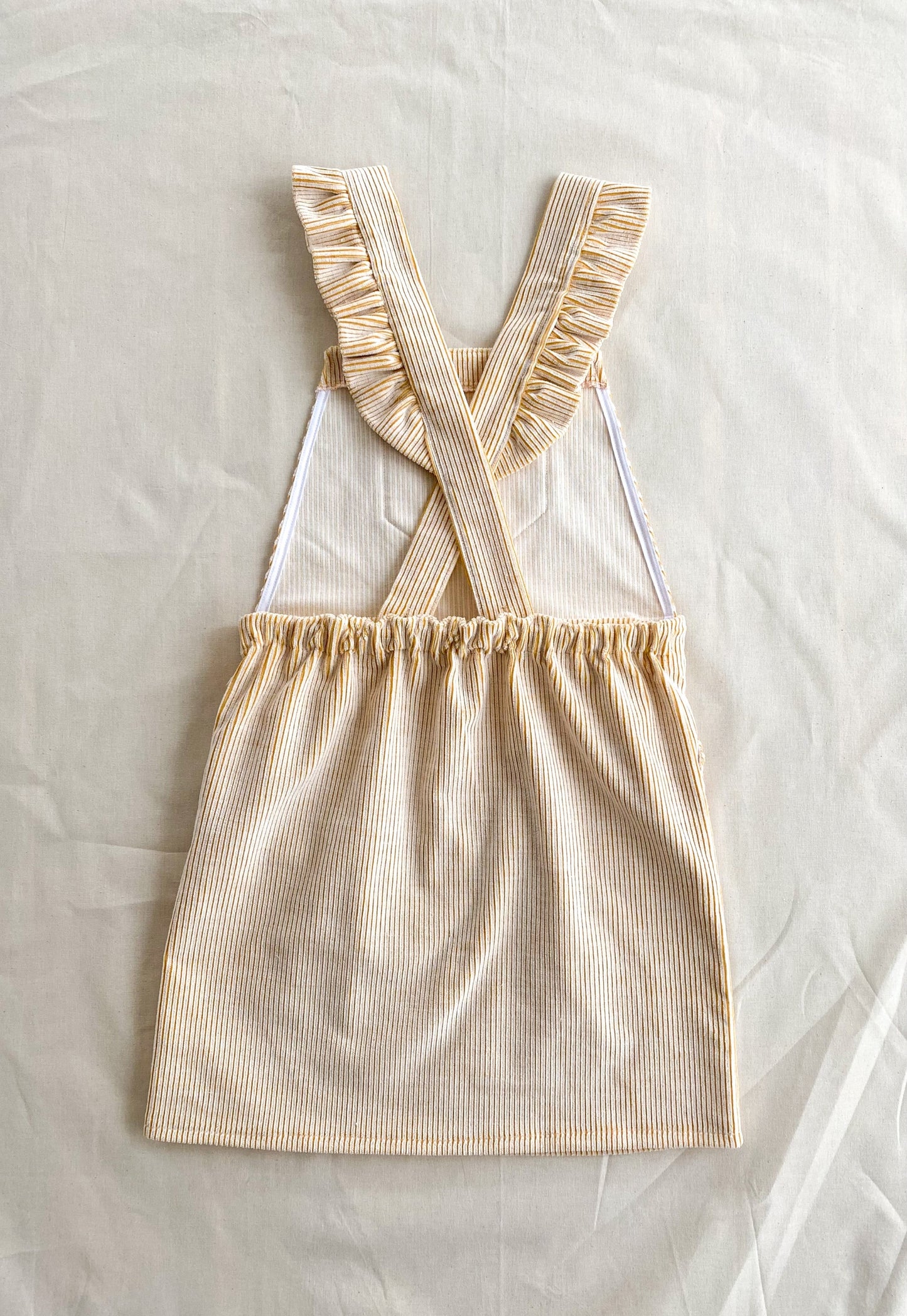 Picture of the Billie pinafore, a short length dungaree dress made from yellow and white deadstock corduroy. The picture shows the back of the dress which is finished with ruffles on the shoulder straps and an elastic in the waistband.