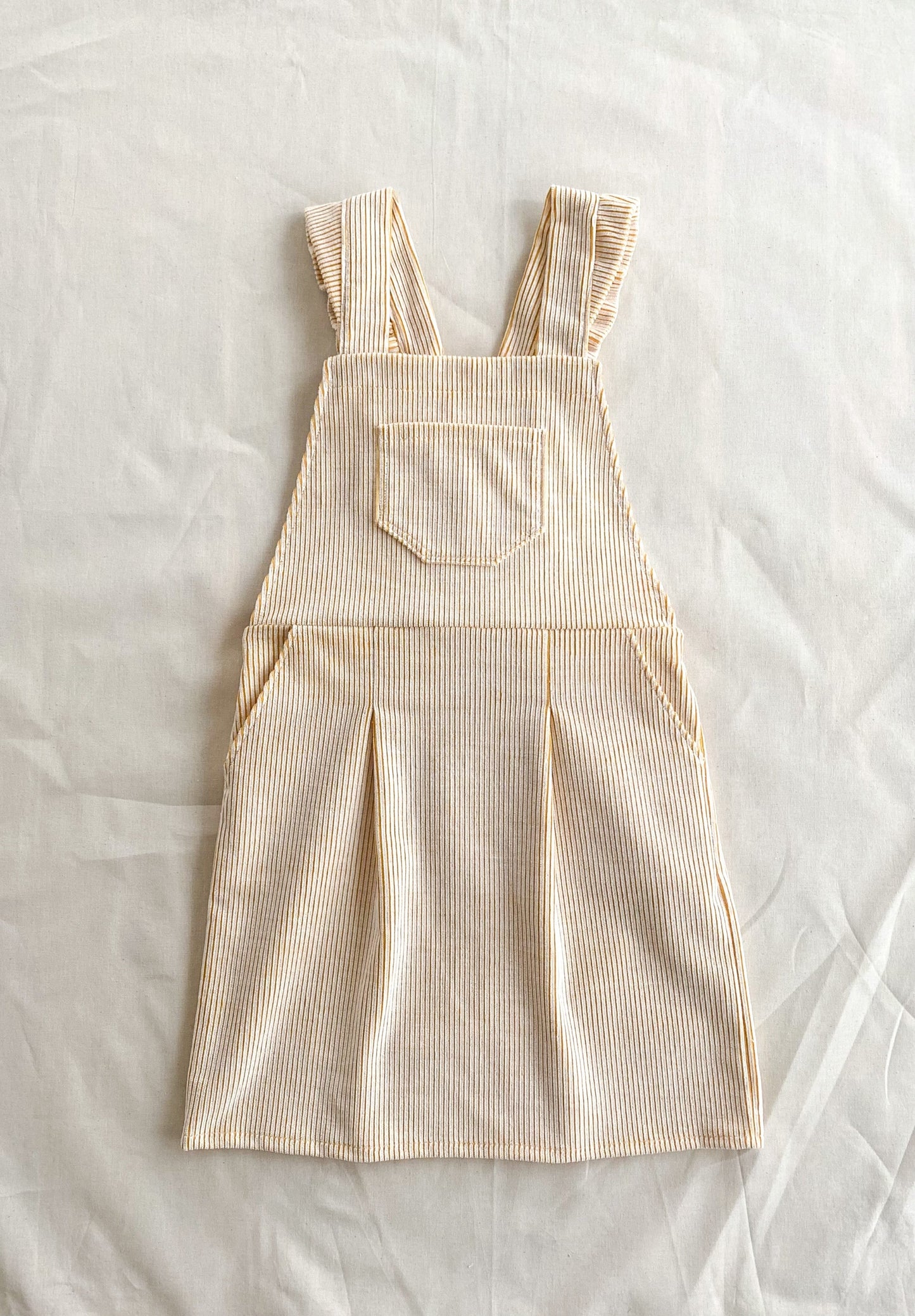 Full front picture of the Billie pinafore, a short length dungaree dress made from yellow and white deadstock corduroy. The dress has pockets in the skirt and a pocket in the top front part.