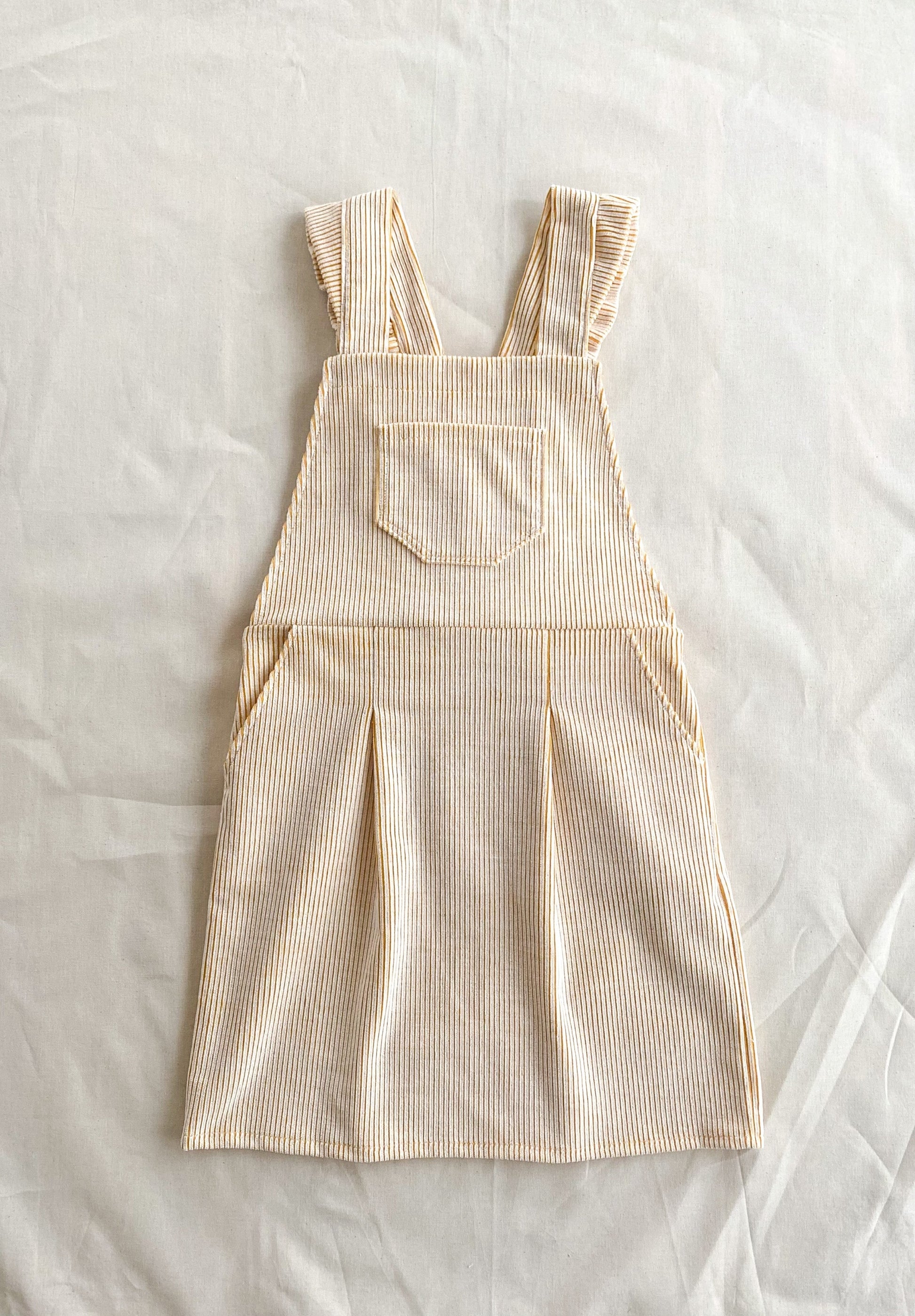 Full front picture of the Billie pinafore, a short length dungaree dress made from yellow and white deadstock corduroy. The dress has pockets in the skirt and a pocket in the top front part.