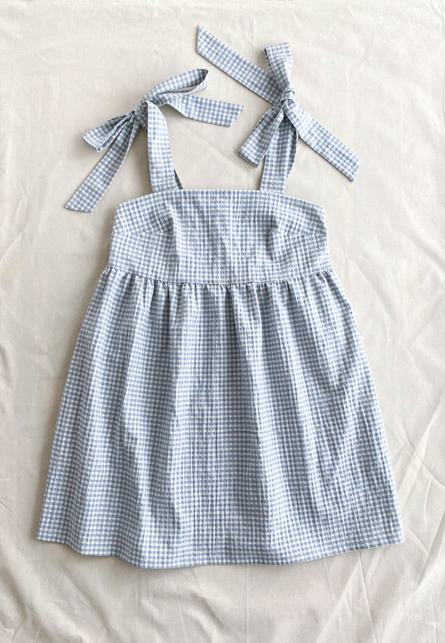 Picture of the front of the Alice dress, a poofy midi length dress made from lightweight deadstock cotton in a blue and white gingham print. The dress is sleeveless and ties into bows at the shoulders.