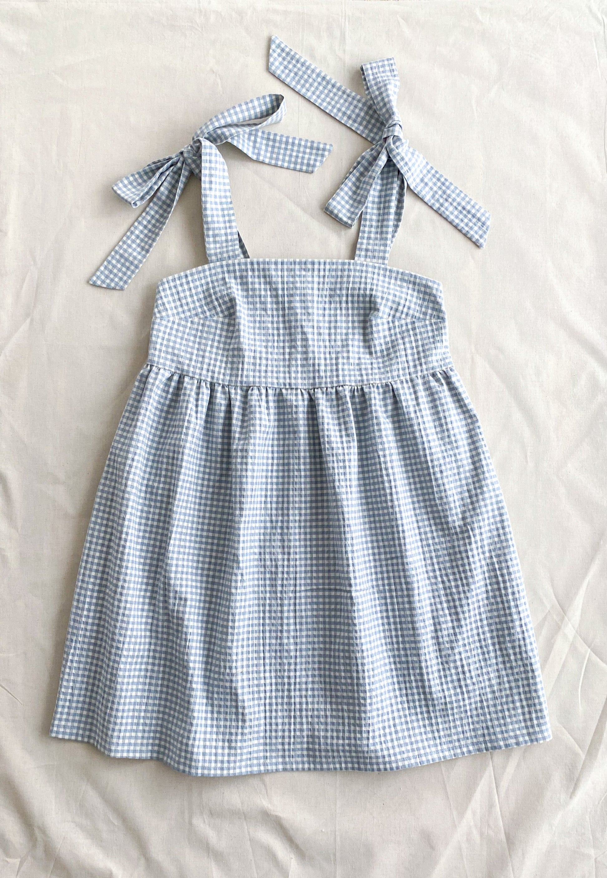 Picture of the front of the Alice dress, a poofy midi length dress made from lightweight deadstock cotton in a blue and white gingham print. The dress is sleeveless and ties into bows at the shoulders.
