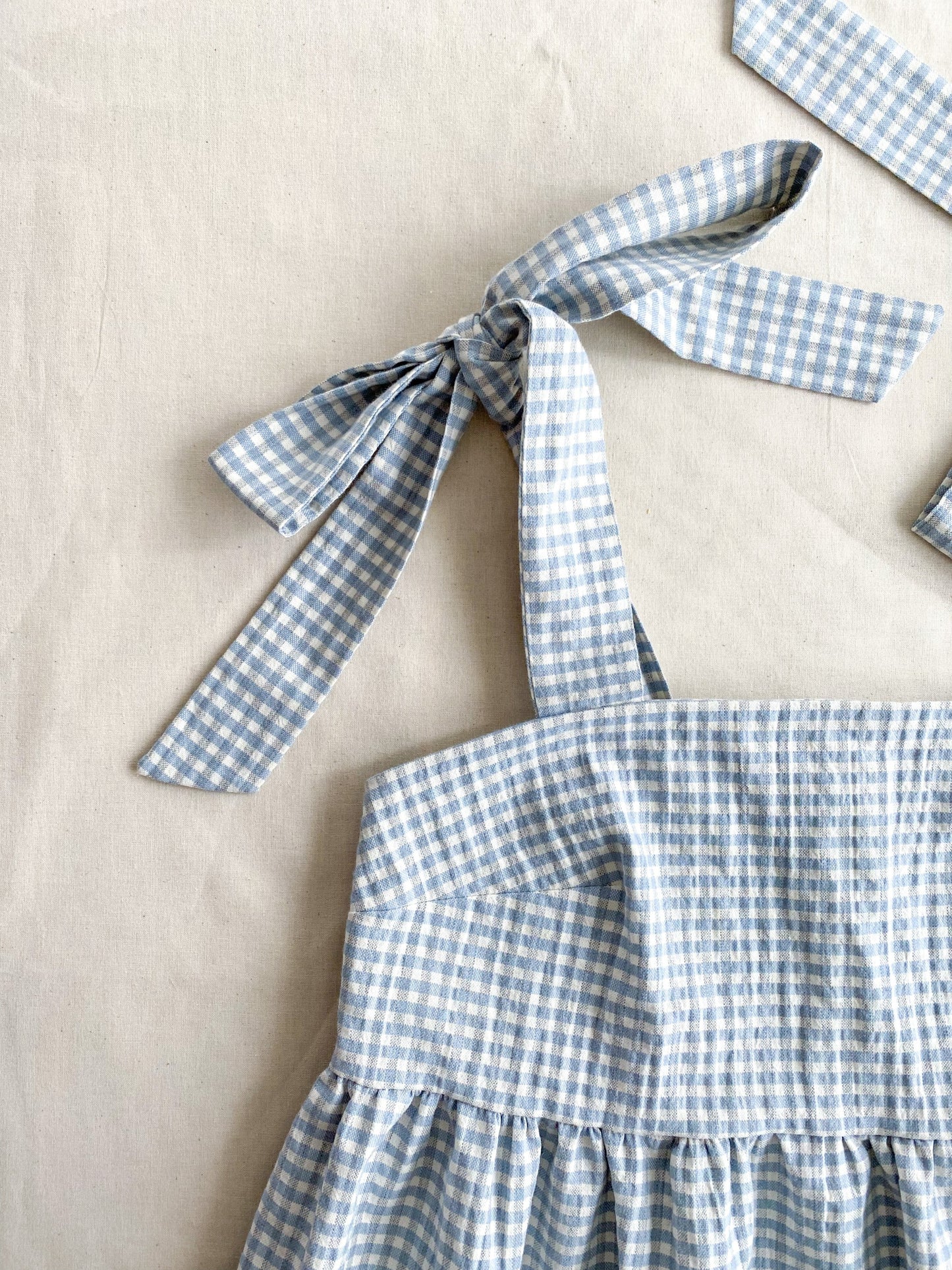Close-up of the Alice dress, a poofy midi length dress made from lightweight deadstock cotton in a blue and white gingham print. The dress is sleeveless and ties into bows at the shoulders.