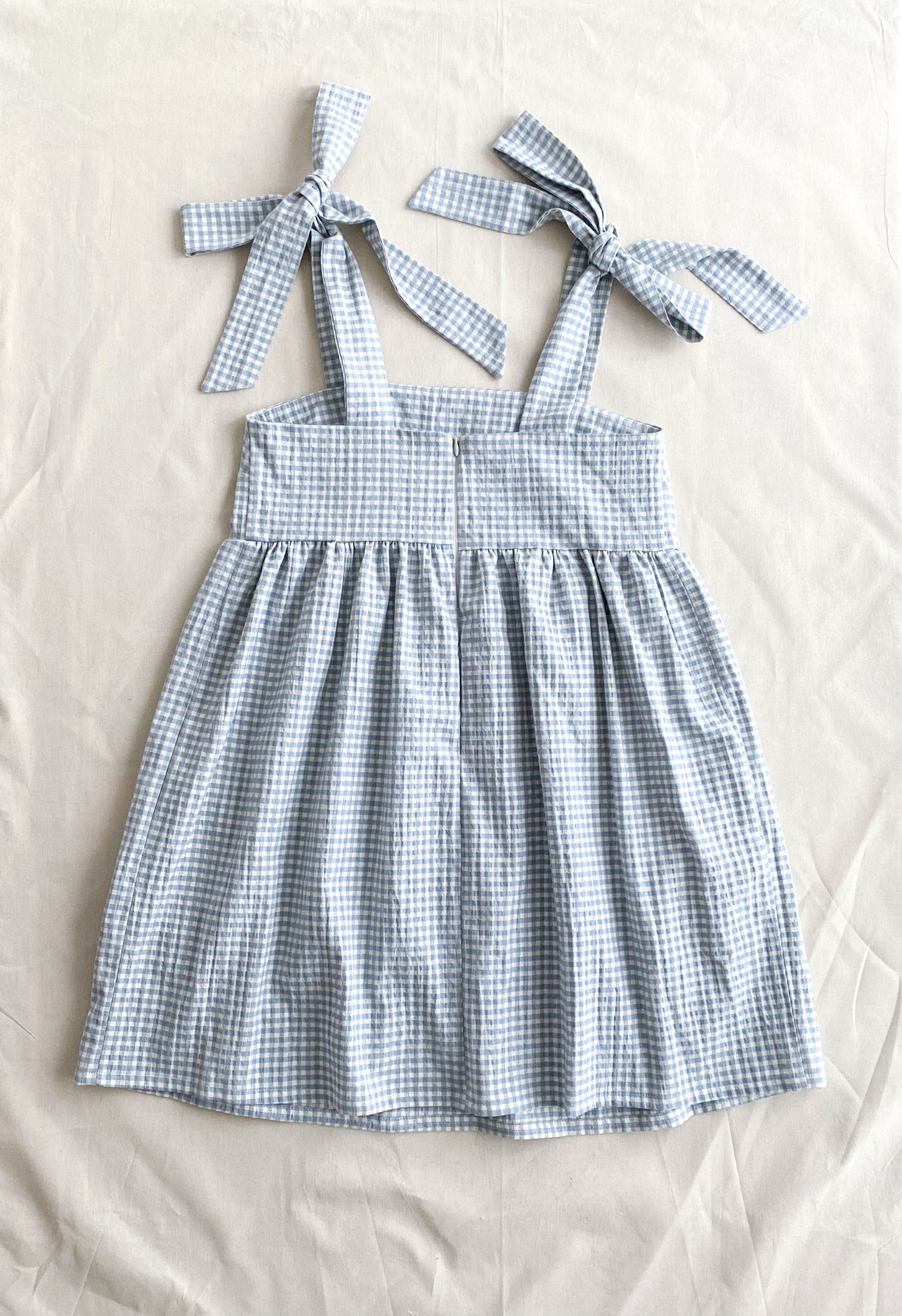 Picture of the back of the Alice dress, a poofy midi length dress made from lightweight deadstock cotton in a blue and white gingham print. The dress is sleeveless and ties into bows at the shoulders. The back closes with an invisible zipper.