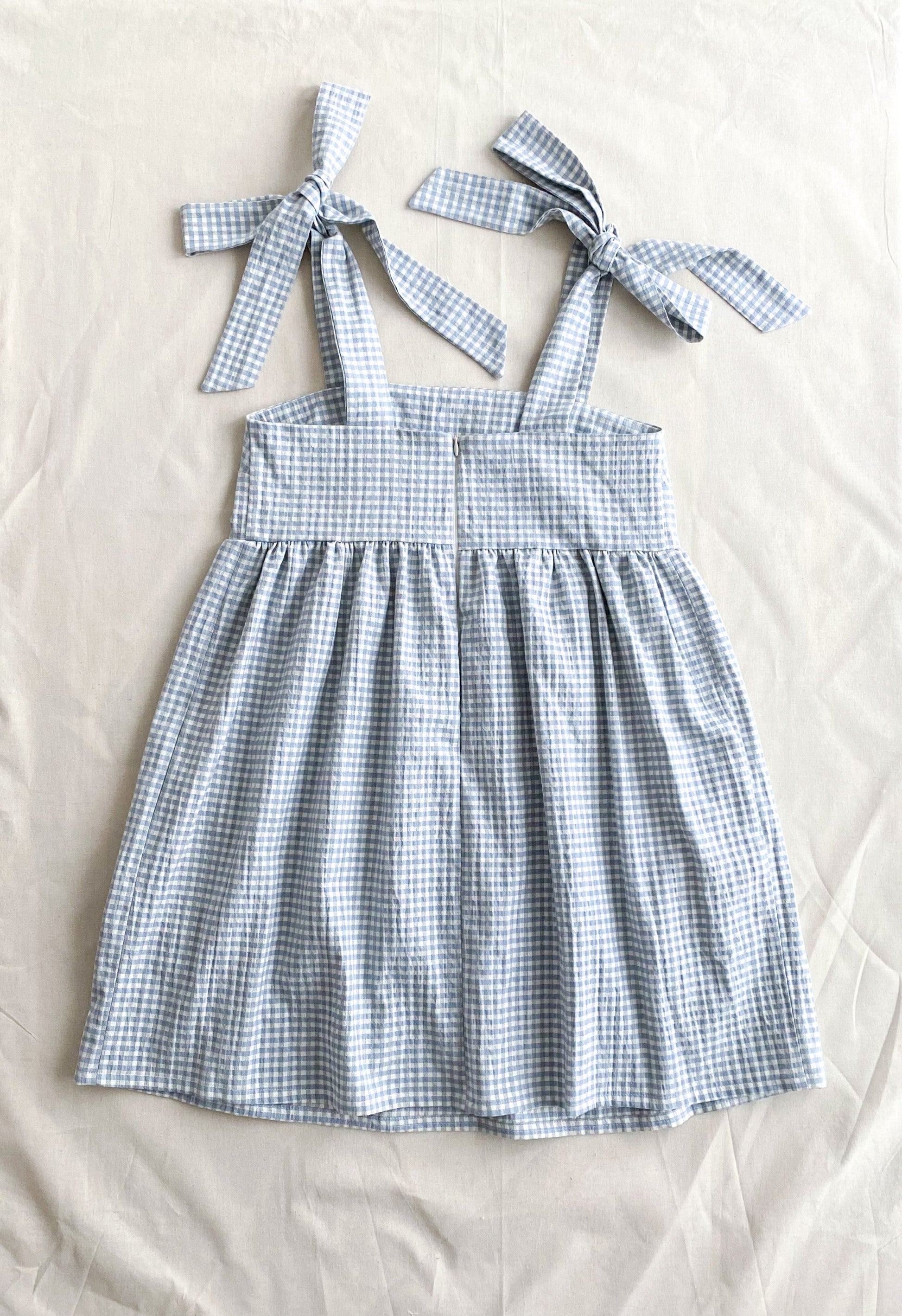 Picture of the back of the Alice dress, a poofy midi length dress made from lightweight deadstock cotton in a blue and white gingham print. The dress is sleeveless and ties into bows at the shoulders. The back closes with an invisible zipper.