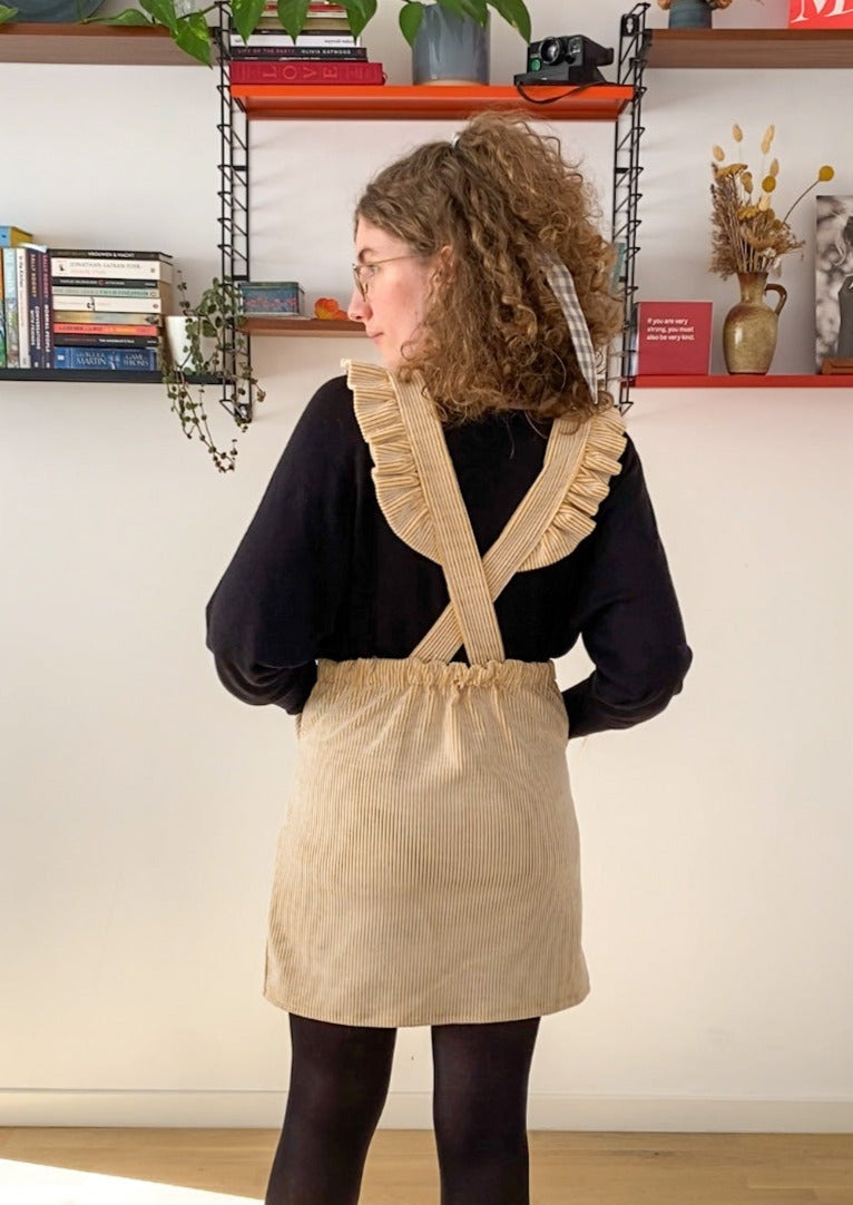 Picture of Leen, a twenty something white woman, posing in the Billie pinafore, showing the back of the pinafore which is finished with ruffles.