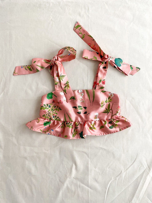 Picture of the front of the Meadow top, a cropped top made from a pink deadstock silk/cotton blend in a beautiful floral print.  The top is sleeveless and ties into bows at the shoulders. The bottom is finished with a ruffle.